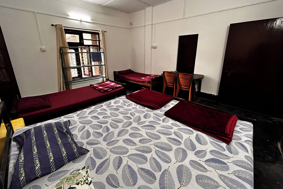 A room with two single beds, a desk with chairs, a window with curtains, and a ceiling fan.