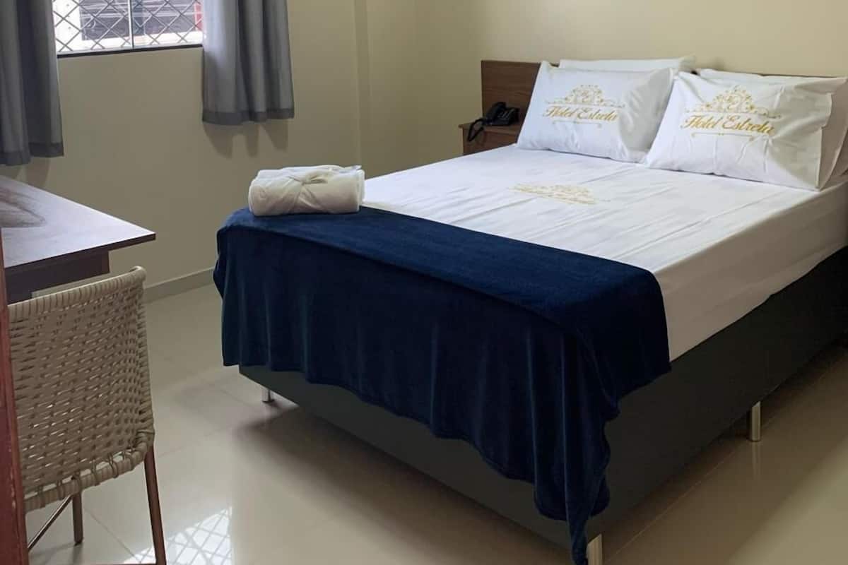 A hotel room with a bed, a desk, an air conditioner, a window with curtains, and a painting on the wall.