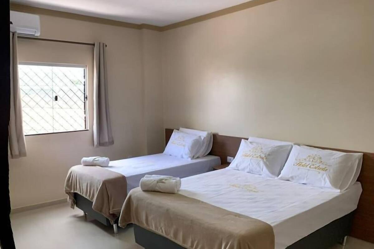A hotel room with two beds, a window with curtains, a wooden table, and a tiled floor.