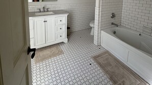 Un baño con suelo de baldosas blancas hexagonales, un tocador blanco con espejo y una bañera con cabina de ducha.