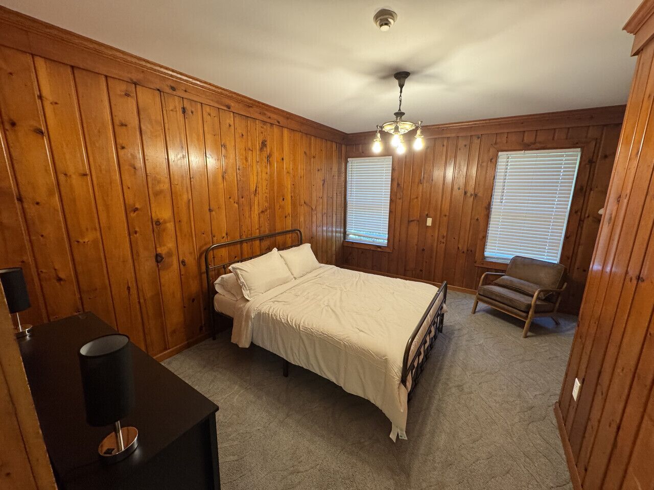 A bedroom with wooden paneling, a bed, a chair, a desk with a lamp, and a ceiling fan.