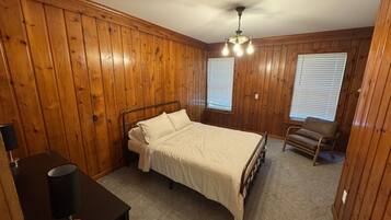 A bedroom with wooden paneling, a bed, a chair, a desk with a lamp, and a ceiling fan.