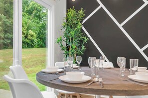 A dining table set for six with white plates, glasses, and cutlery, a potted plant, and a view of a grassy area through large windows.