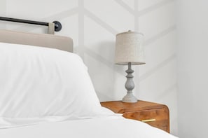 A bed with white bedding, a wooden nightstand with a lamp, and a geometric-patterned wall. - The Spartan Retreat | Luxe 3BR | Sleeps 10 (Spartanburg)
