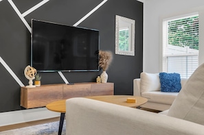 A modern living room with a flat-screen TV mounted on a wall, a wooden console table, a white sofa with a blue pillow, and a window with blinds. - The Spartan Retreat | Luxe 3BR | Sleeps 10 (Spartanburg)