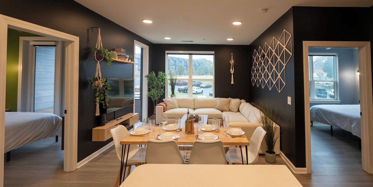 A modern living room with a sofa, dining table, and chairs. There are two beds in the background.