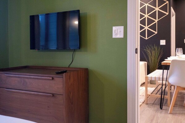 A room with a flat-screen TV mounted on the wall, a wooden cabinet with drawers, and a dining area with a table and chairs. - Luxe Modern Loft, Great for Long Stays W/King Beds (Spartanburg)