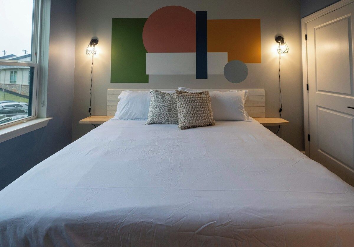 A neatly made bed with white linens and two decorative pillows, a wall with abstract art, and two wall-mounted lamps on either side.