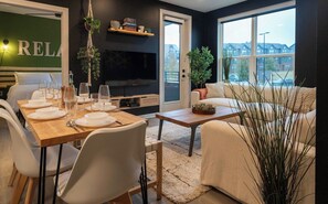 A modern living room with a dining area, a flat-screen TV, and a large window offering a view of the outside. - Luxe Modern Loft, Great for Long Stays W/King Beds (Spartanburg)