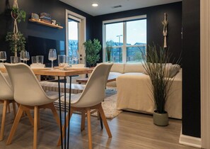 A dining area with a table set for four, surrounded by white chairs. There is a sofa and a potted plant in the background.