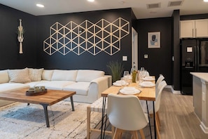 A modern living room with a white sofa, a wooden coffee table, and a dining area with a wooden table set for four.