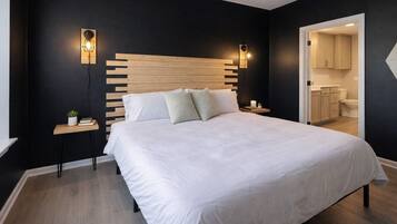 Un dormitorio con cabecera de madera, ropa de cama blanca, dos mesitas de noche con lámparas y vista al baño.