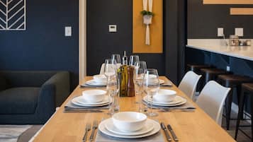 Mesa de jantar posta com pratos brancos, talheres e copos de vinho, com uma planta e uma luminária na parede ao fundo.