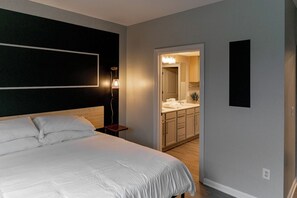 A bedroom with a large bed, a bedside table, a wall-mounted light, and a view into a bathroom with a sink and mirror. - Ideal for long stays, great location, King bed! (Spartanburg)
