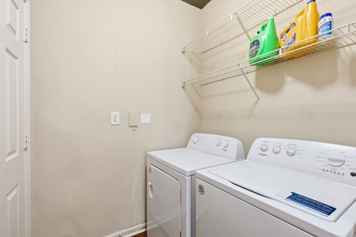 Un área de lavandería con dos lavadoras y secadoras apiladas, un estante con productos de limpieza y tomas de corriente en la pared.