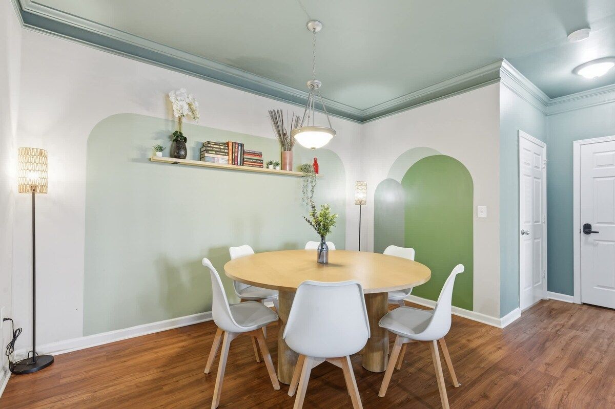 A dining area with a round table, white chairs, a hanging light fixture, and a bookshelf with books and decorative items.