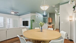 A modern living room with a round wooden table, white chairs, a ceiling fan, and a flat-screen TV.