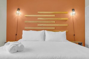 A hotel room with a bed, white linens, a wooden headboard, and two wall-mounted light fixtures. - Pool, Gym & Central Location! 2- King Beds (Spartanburg)