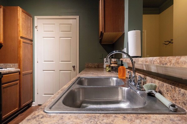 A kitchen with a double sink, wooden cabinets, a white door, and a countertop with a paper towel holder. - 3 King Beds, Perfect for Long Stays (Spartanburg)