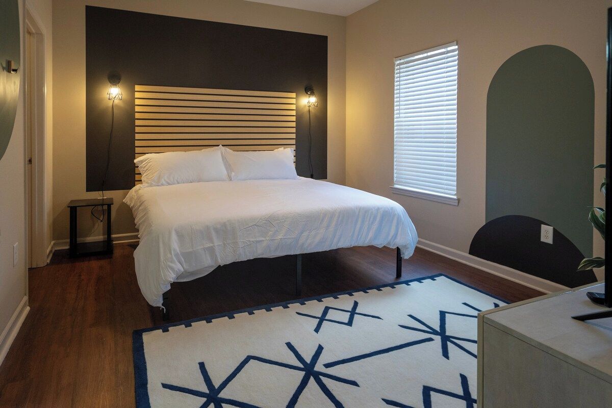 A bedroom with a bed, bedside lamps, a window with blinds, a rug, and a desk with a chair.