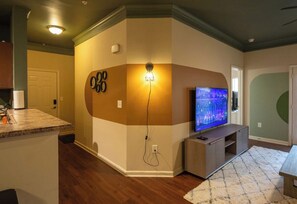 A living room with a television, a wall-mounted light, and a kitchen area in the background. - 3 King Beds, Perfect for Long Stays (Spartanburg)