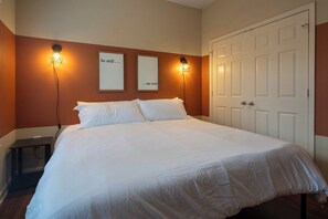 A neatly made bed with white linens in a room with orange walls and a white door.