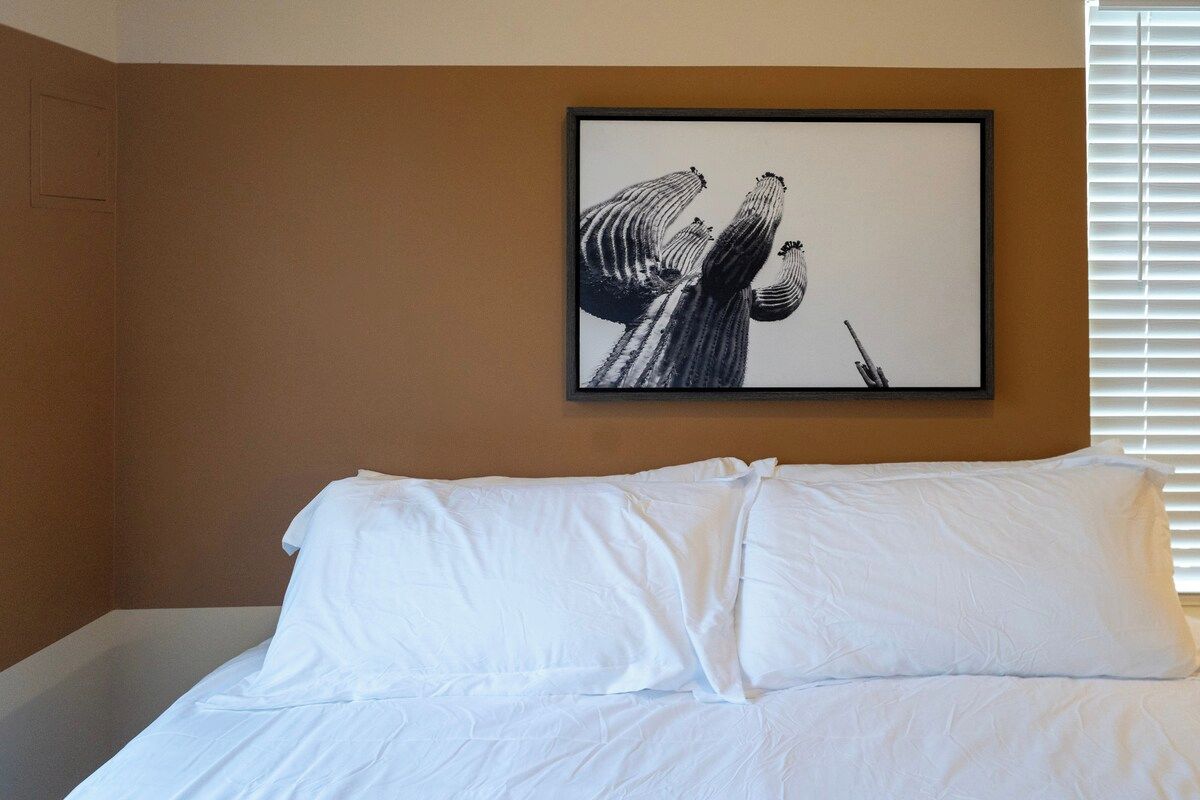 Quarto de hotel com uma cama, uma obra de arte emoldurada na parede e uma janela com persianas.