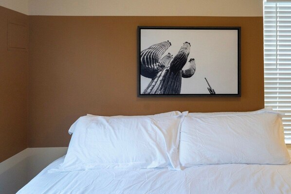 Quarto de hotel com uma cama, uma obra de arte emoldurada na parede e uma janela com persianas.