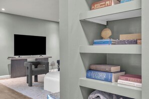 Ein modernes Wohnzimmer mit einem Flachbildfernseher, einem grauen Sofa und einem Bücherregal mit Büchern und Dekorationsobjekten.