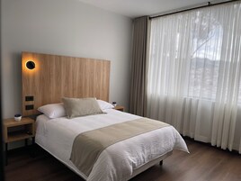 Habitación de hotel moderna con cabecera de madera, cama grande con manta color beige, mesita de noche con lámpara y cortinas traslúcidas sobre una ventana.
