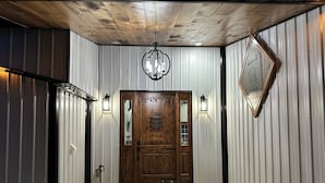 A wooden door with a glass panel, flanked by two wall-mounted lights, leading to a room with a wooden ceiling and a metal wall. - Rippling Waters! 3 BD 2.5 BA Riverfront Home, 8 min. from airport.  Sleeps 11. (Mills River)