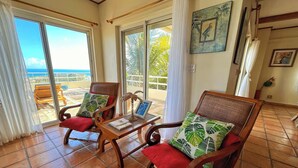 A room with two wooden chairs with red cushions, a small table with a book and a framed picture, and a view of the ocean through large sliding glass doors.