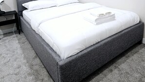 Een moderne slaapkamer met een grijs gestoffeerd hoofdeinde, wit bedlinnen en een opgemaakt bed.