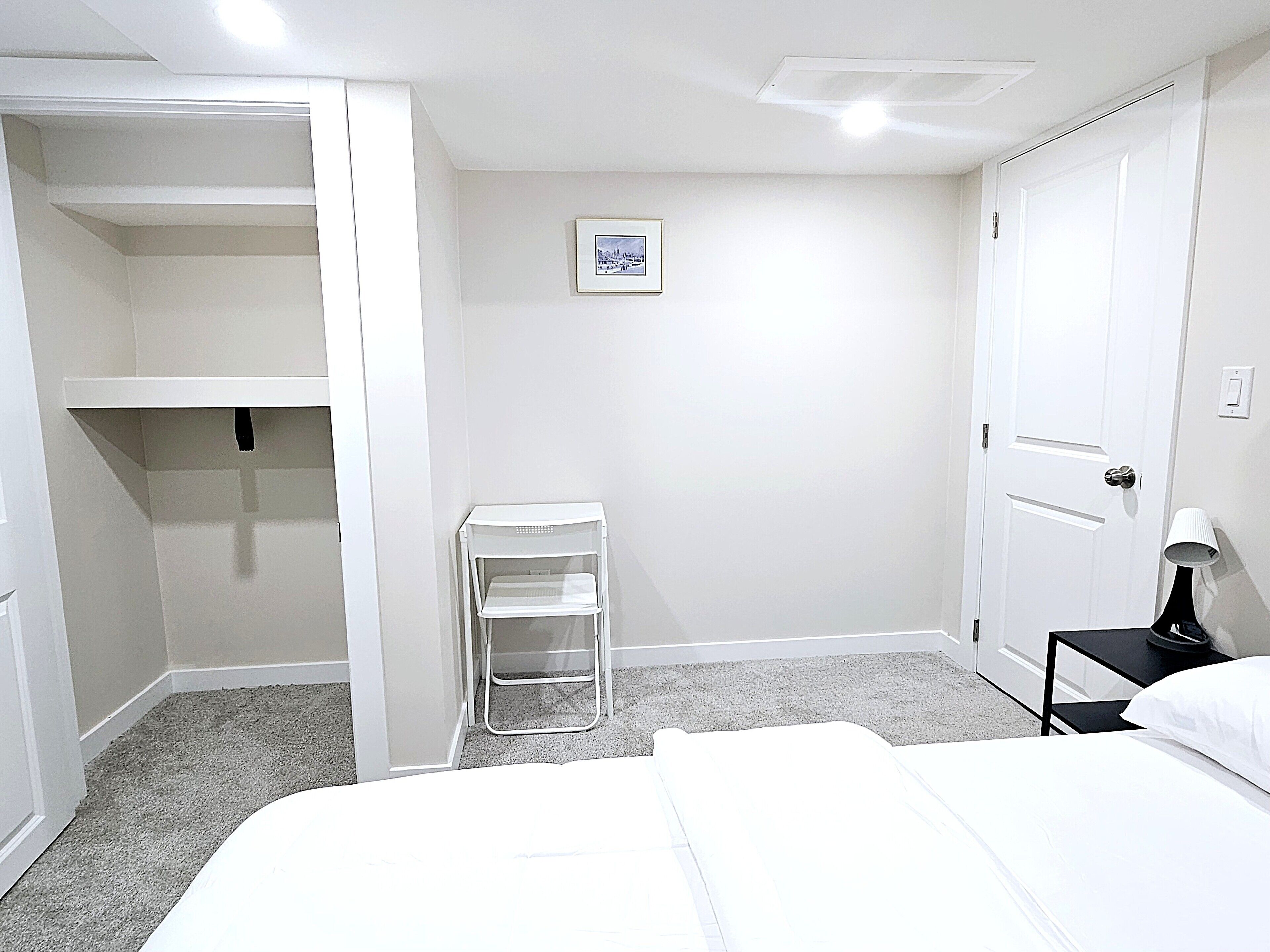 Une chambre d’hôtel avec un lit, une armoire blanche, une petite table blanche, une table de chevet avec une lampe et un tableau encadré au mur.