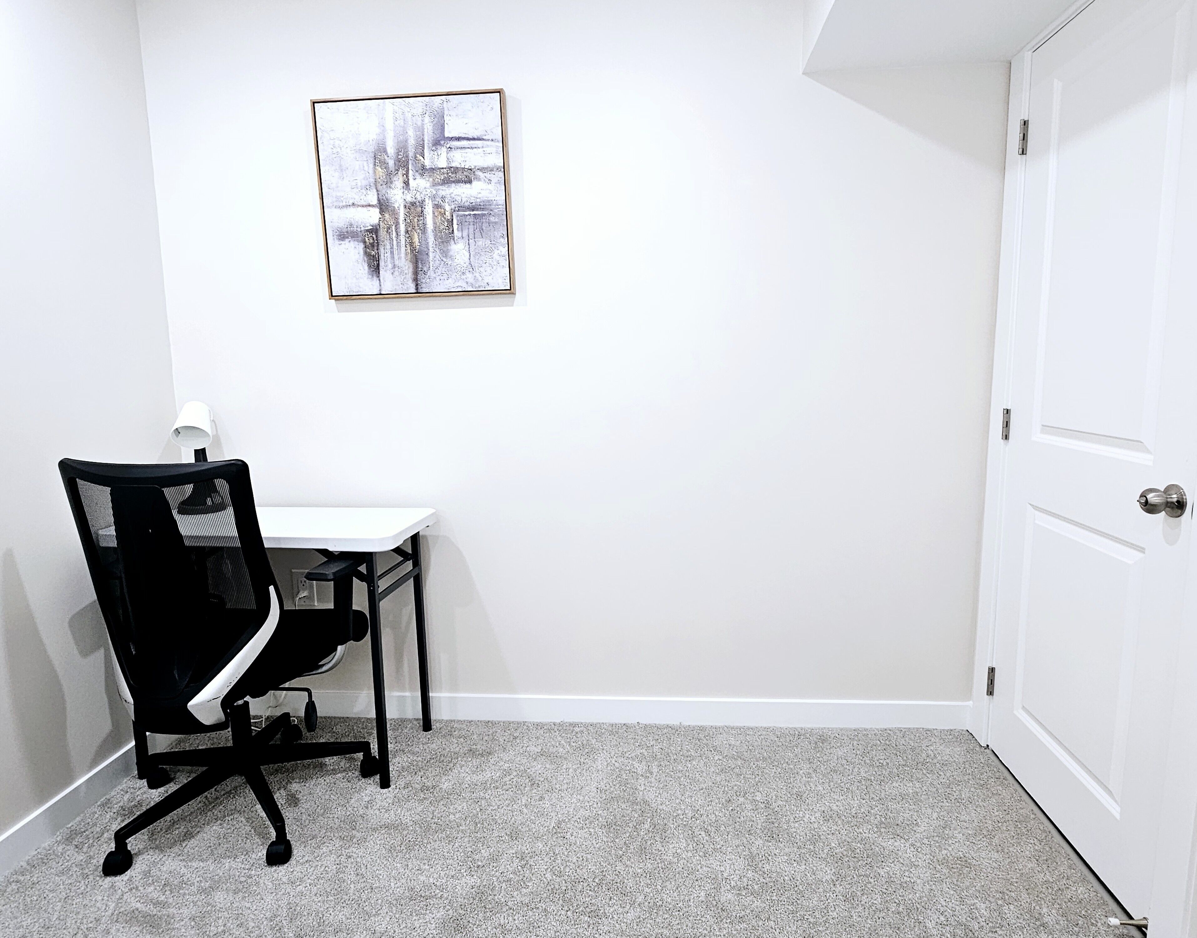 Un petit bureau avec un bureau blanc, une chaise de bureau noire, un tableau abstrait encadré accroché au mur et une porte blanche.