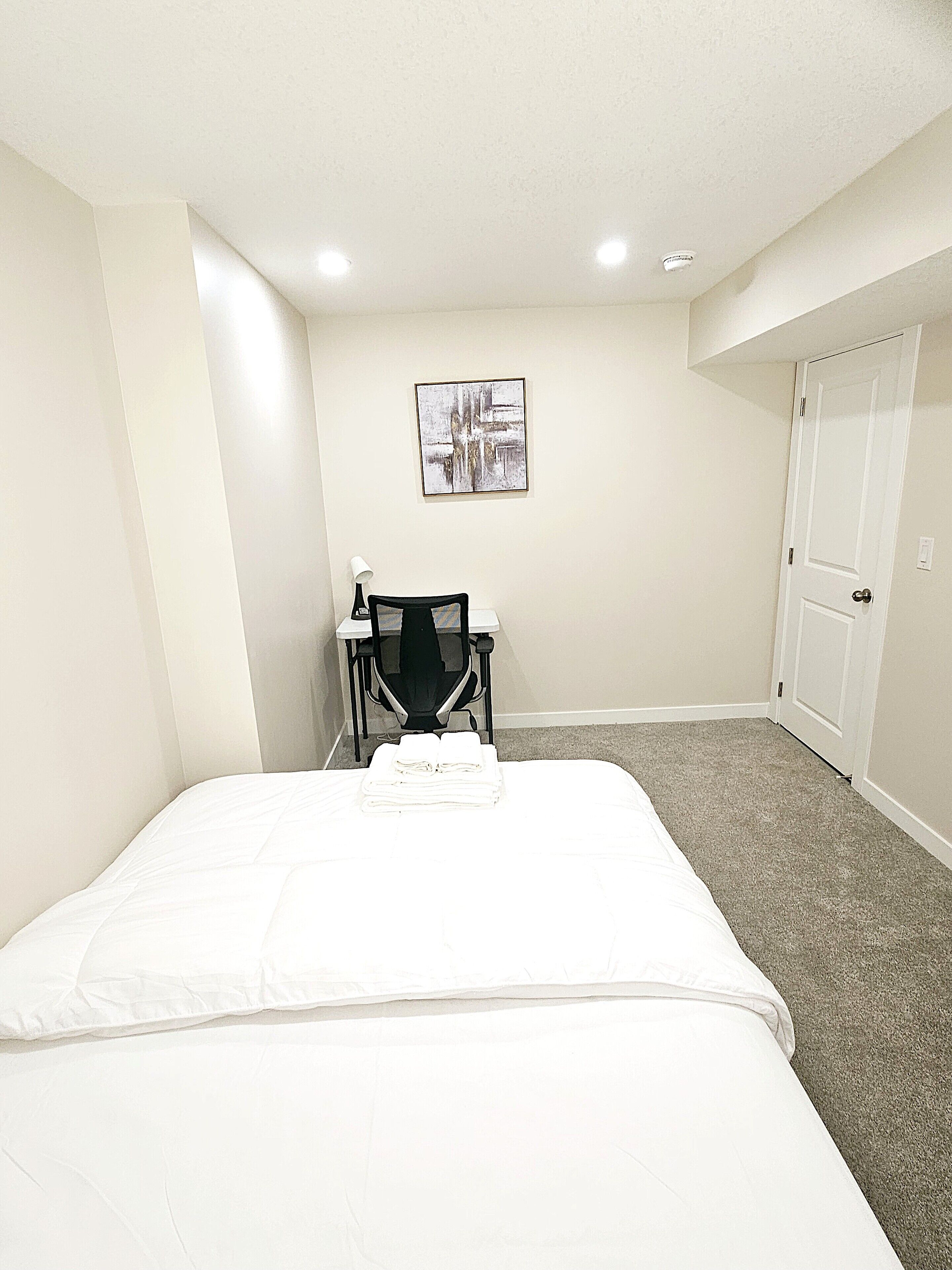 A hotel room with a neatly made bed, a chair, a desk with a lamp, a painting on the wall, and a closet.