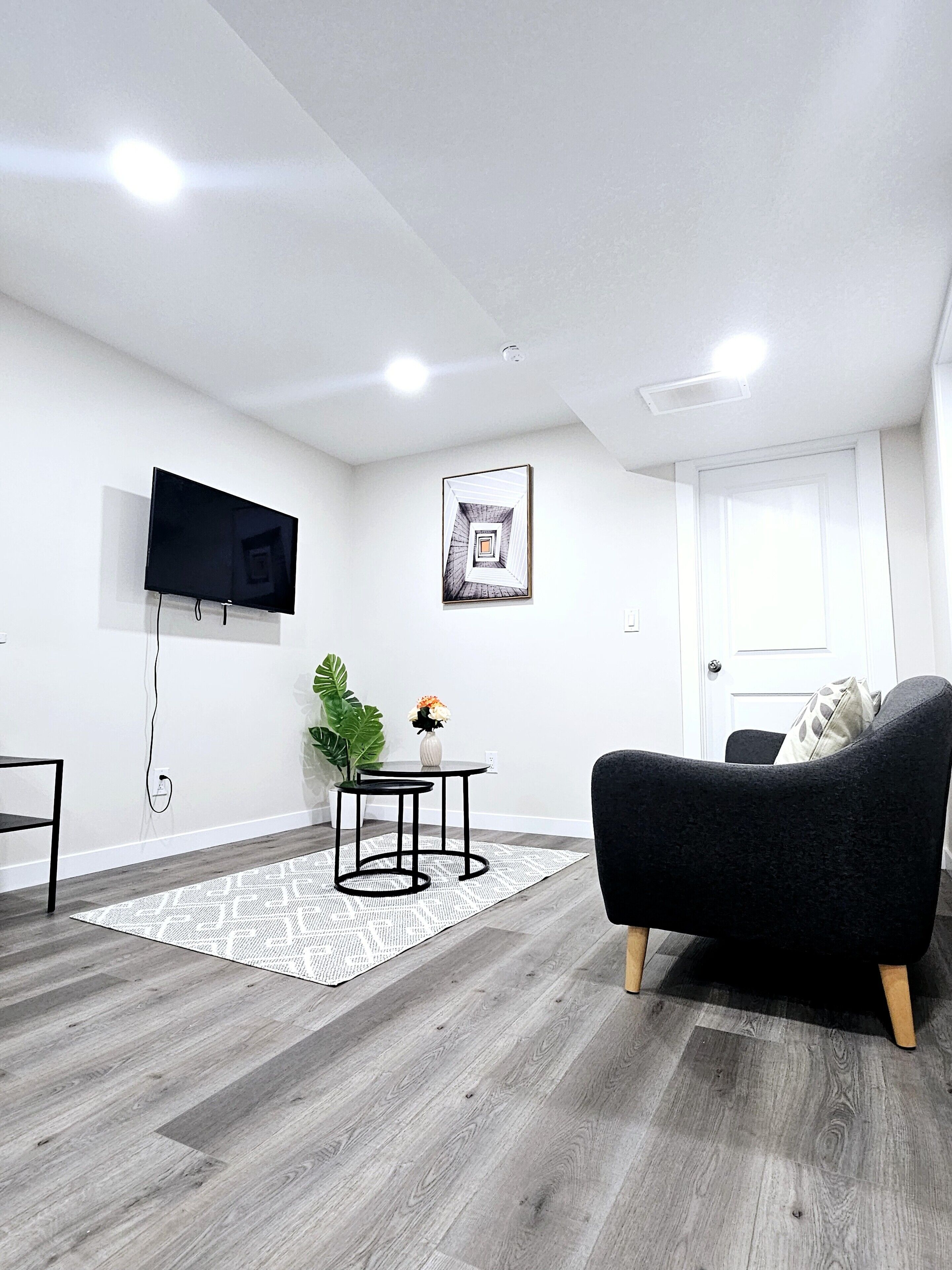Un salón moderno con un televisor de pantalla plana, una mesita redonda con dos sillas, un sofá negro y una planta en maceta.