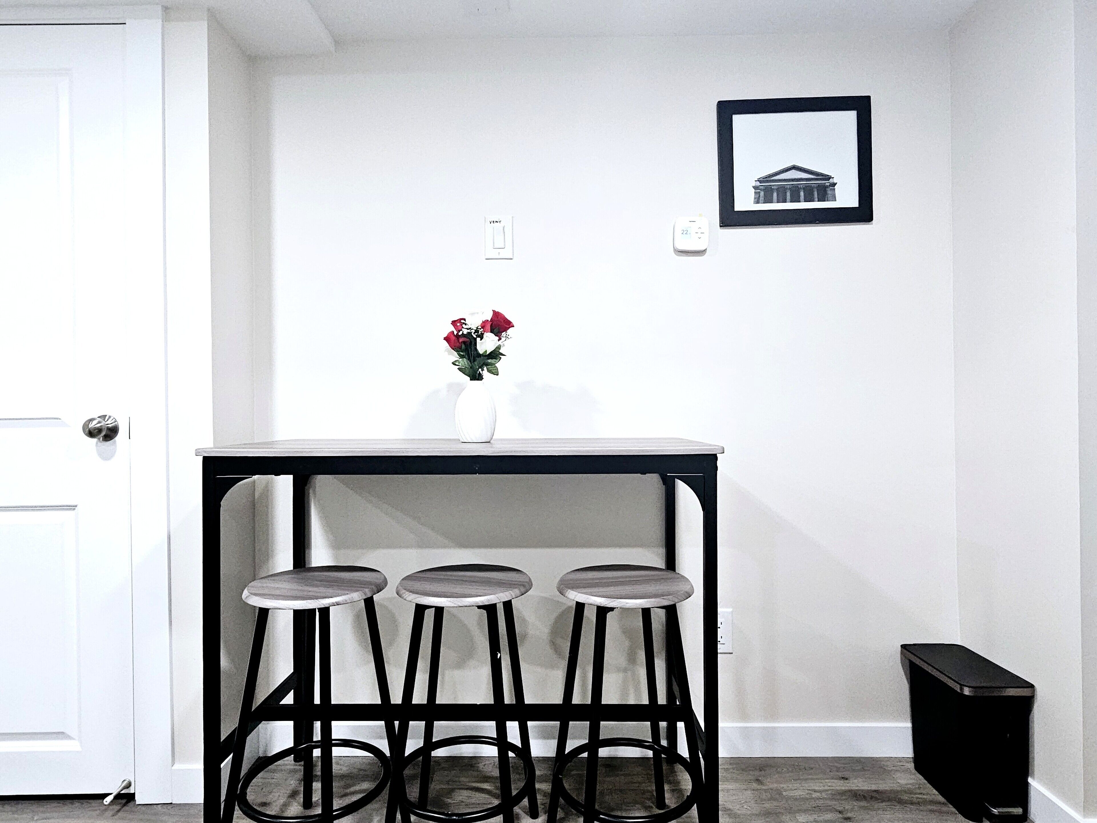 A small kitchen area with a white door, a black table with three stools, a vase with flowers, and a framed picture on the wall.
