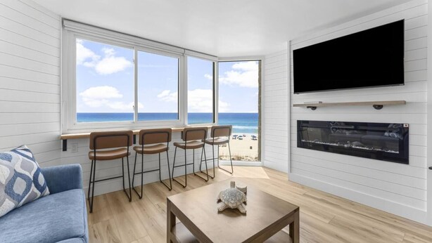 A room with a large window overlooking a beach, a flat-screen TV mounted on the wall, and a small table with a turtle figurine. - Beach Front Ocean View Condo - Top of the Gulf 521 (Panama City Beach)
