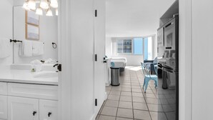 A modern kitchen with white cabinets, a sink, and a trash can. There is a window with blue curtains and a chair in the background.
