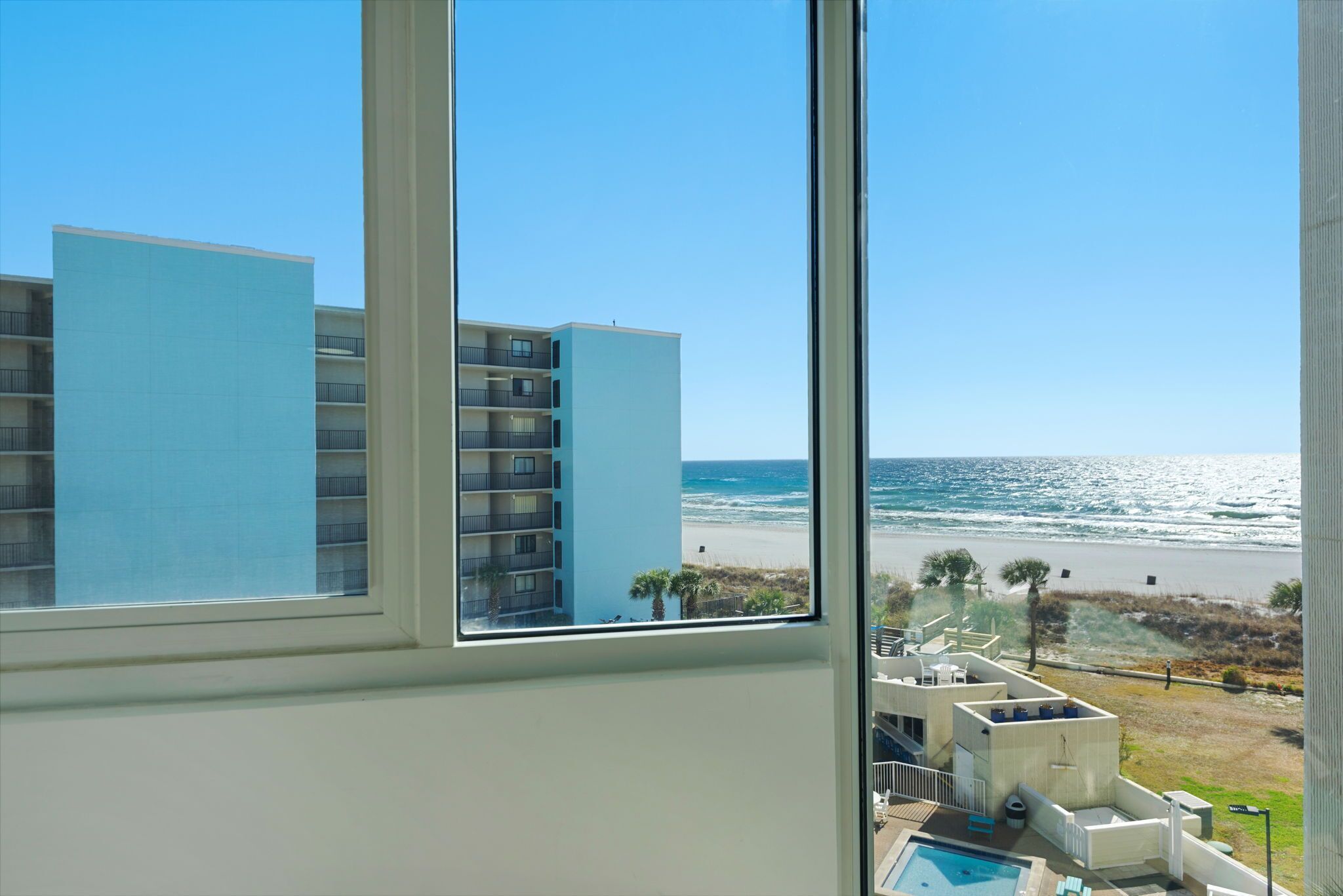 Une vue depuis une fenêtre donnant sur une plage, avec une piscine et un bâtiment en arrière-plan.