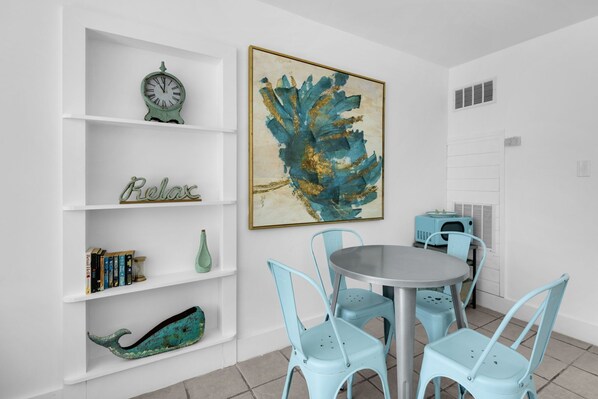 A dining area with a round table and four light blue chairs, a white bookshelf with various items, and a large abstract painting on the wall.