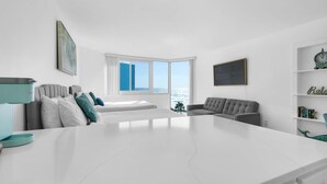 A modern living room with a large white table, grey sofas, a bookshelf, and a view of the sea through large windows.