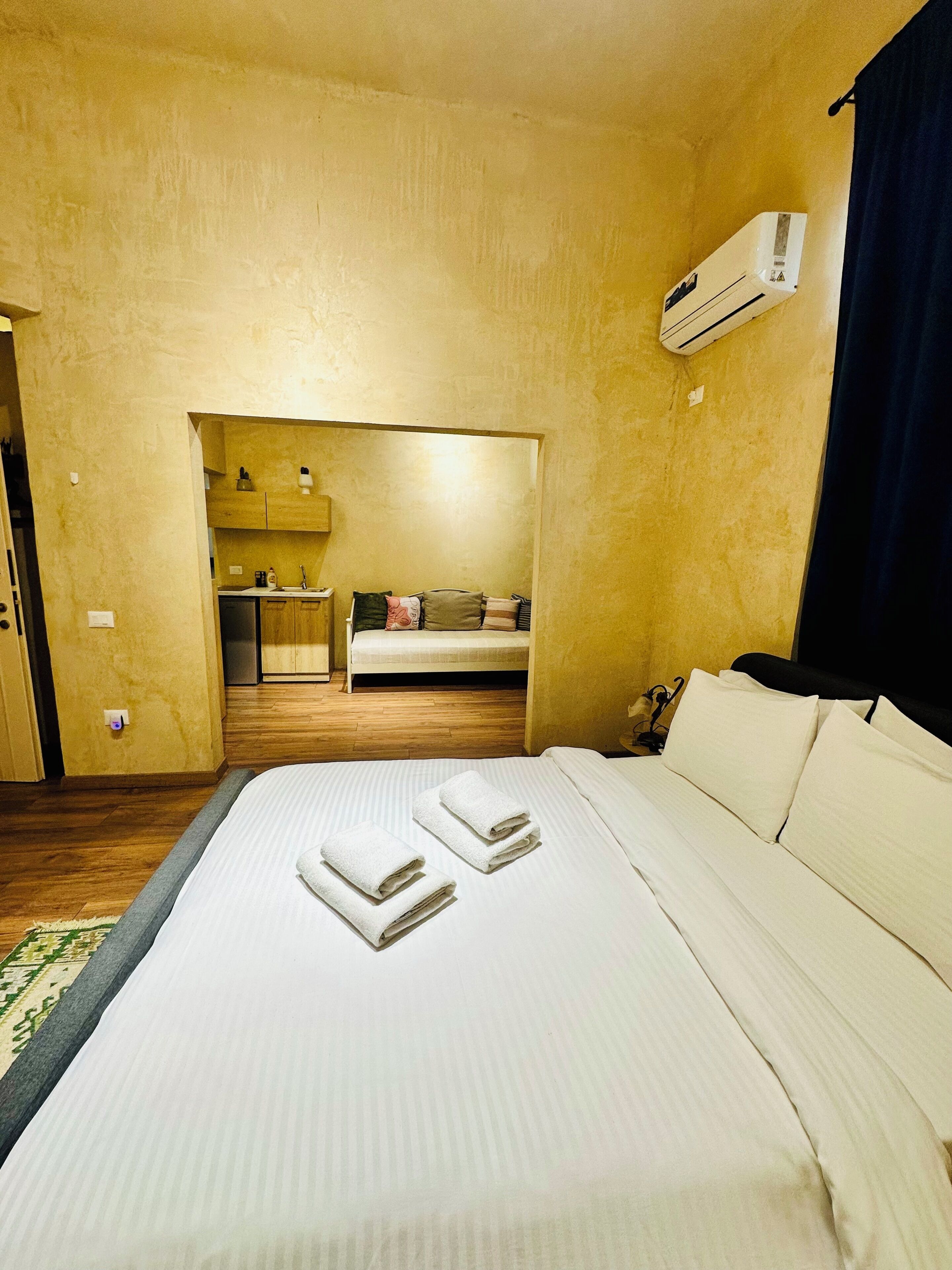 A hotel room with a bed, a small kitchenette, a sofa, and an air conditioning unit.