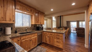 A spacious kitchen with wooden cabinets, granite countertops, and a central island. There is a dining area with a table and chairs, and a view of the outdoors through large windows. - Charming and Relaxing Getaway with Great Views (Shaver Lake)