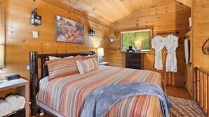 A wooden cabin bedroom with a bed, a nightstand, a painting, and a window with a view of greenery.