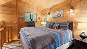 A wooden cabin bedroom with a bed, nightstands, a desk, and a window with blinds.