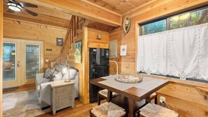 A cozy cabin interior with a dining area, a sofa, a staircase, and a window with curtains.