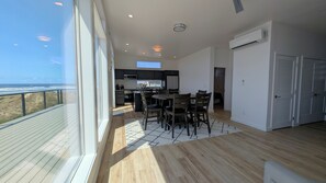 A modern kitchen with dark cabinets, a dining area with wooden chairs, and a view of the ocean through large windows.