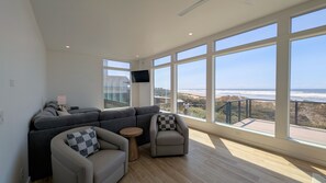 Un vaste espace de vie avec une grande fenêtre donnant sur une plage, un canapé d’angle gris, une petite table ronde et un fauteuil gris.